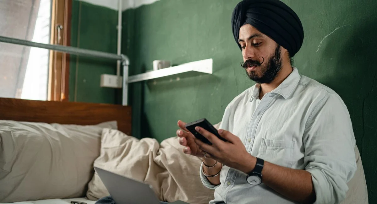 Concentrated bearded Sikh male wearing turban sitting on bed with pillows in green room near window with netbook and browsing